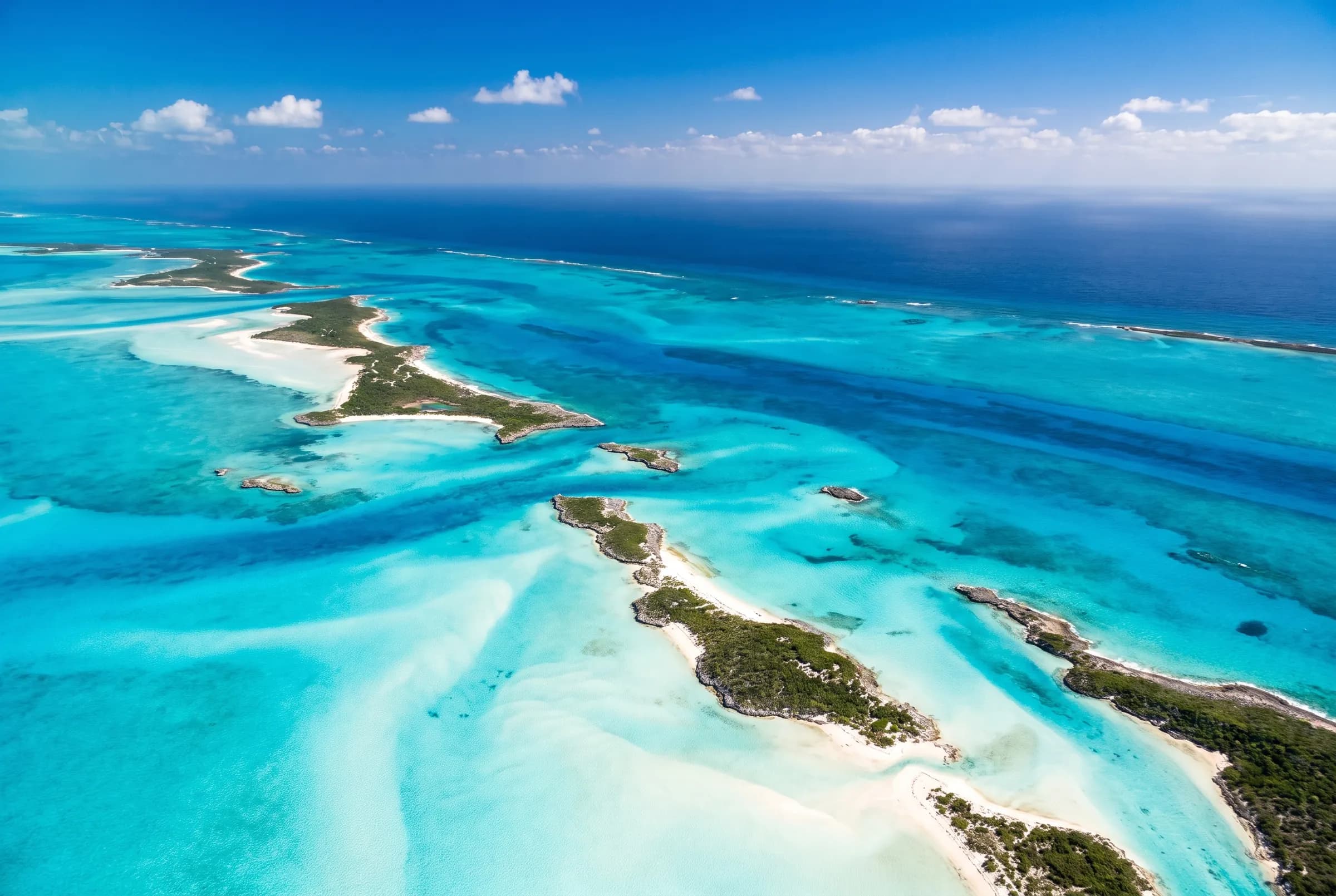 Aerial view of the Exuma cays in The Bahamas — green limestone islands and white sandbars scattered through turquoise shallows, with deep cobalt channels running between them.