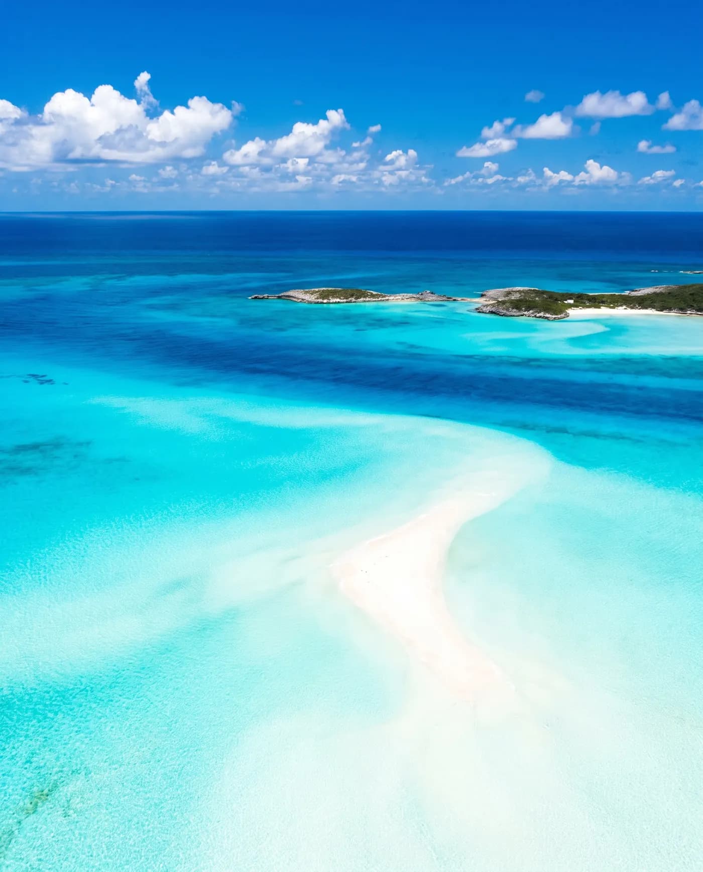 Aerial view of a white sandbar curving through brilliant turquoise shallows in The Bahamas, with small green cays and deep blue ocean beyond.